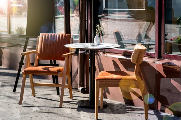 a round table with two chairs with backs on the wooden legs of a summer cafe on the sidewalk near the facade of the building with panoramic windows of dark color with a sun flare.