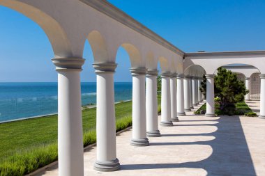 sütunlu kemerli binalar ve çam ağaçları ile bir çiçek yatak ve arka planda mavi bir gökyüzü ile bir deniz ufku ile taş çini mermer bir zemin ile evin arka bahçesinde.