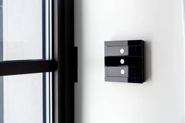 touch glass light switch of black color on a white wall near the window, close-up of the smart home control with 3 buttons.