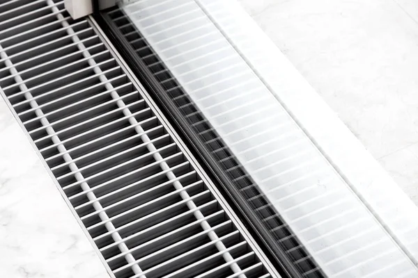 Panoramik pencerenin yanındaki beyaz taş zemin döşemesinde zemin havalandırmalı ısıtma ızgarası, klima ızgarasını kapatın.