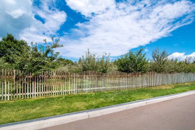 bir kaldırım ve drenaj sistemi ve yeşil çim ile asfalt yol kenarında ahşap bir çit arkasında meyve bahçesi.