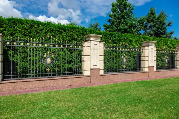 an iron wrought fence with capital stone columns and a sidewalk along a green lawn on a sunny summer day in the backyard.