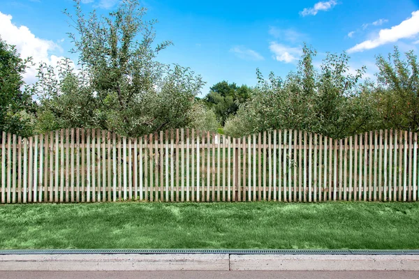 bir kaldırım ve drenaj sistemi ve yeşil çim ile asfalt yol kenarında ahşap bir çit arkasında elma bahçesi.