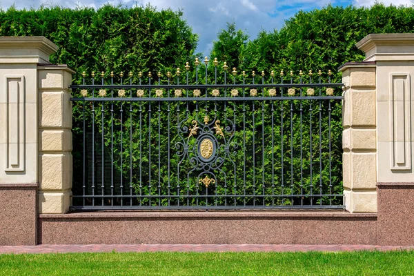 a stone fence with a rustik and an iron forged insert with a pattern and peaks near a thuja hedge and a green lawn with a sidewalk along the fence.