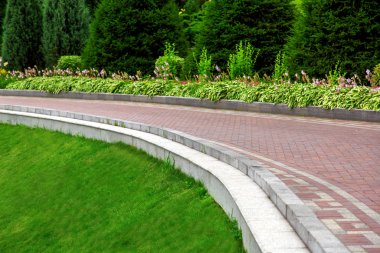 yeşil çimen, çiçek ve çam ağaçları ile bir parkta taş fayans yapılmış bir kaldırım bir yamaç üzerinde bir taş kaldırım ile güçlendirilmiş patika kenarı.