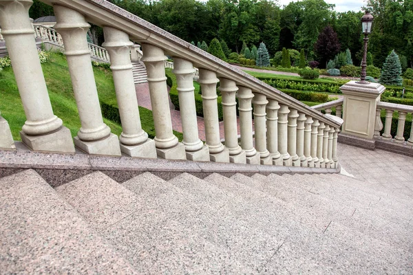 granite staircase with railings and stone balustrades with a pedestal for a street lamp in the background a park with green plants.
