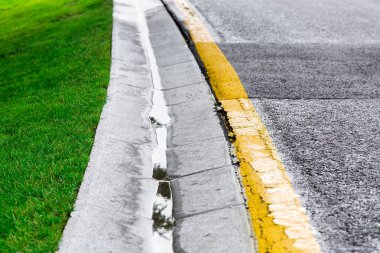 Kanalda akan yağmur suyu, ıslak asfalt yolunun kenarında çimento hendeği ve yol kenarında yeşil bir çimenlik var..