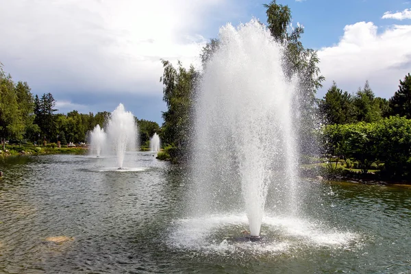 Bulutlu bir gökyüzü ile güneş ışığı ile aydınlatılmış bir yaz parkı bir çeşme su jetleri ile Pond.