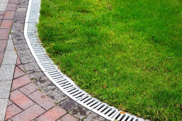 curved iron grid drainage system in the green lawn and footpath of red paving slabs, close up.