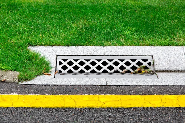 drainage system edge tray with concrete grate for rainwater drainage into the sewer on road with a yellow marking and a green lawn after rain.