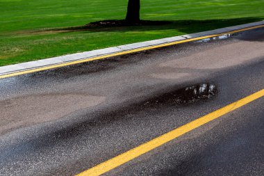 yağmurdan sonra su birikintileri ile ıslak asfalt yol, güneşli bir günde yeşil bir çim tarafında kaldırım yüzeyinde sarı işaretler ile karayolu.