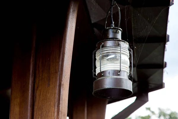 A retro pendant lamp of street lighting made of iron and a glass shade in the web is hanging on a wooden support of a brown gazebo.