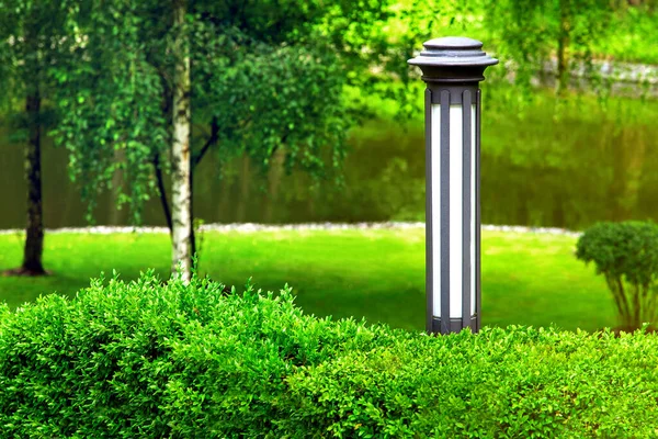 Iron lantern garden lighting near green deciduous bushes in the park with trees and bushes in greenery background in the defocus of a blure pond with water.