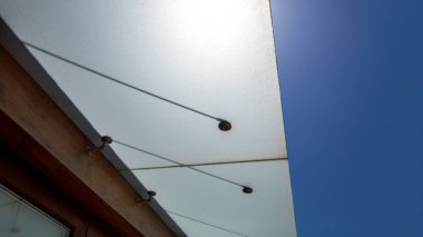 Binanın ön cephesinde cam kaplama güneşlik var. Arka plandaki duvara metal bağlarla sabitlenmiş. Mavi bir gökyüzü ve modern mimarinin detaylarına yakın çekim yapan güneş..