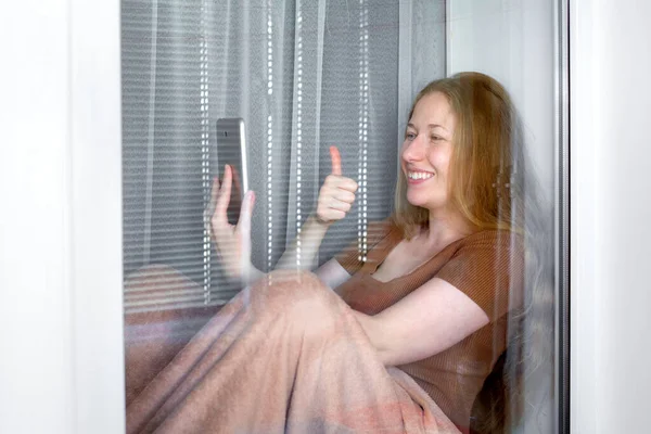 happy smile girl sits on windowsill by window and holding smartphone in one hand with other hand shows gesture class thumb up, working remotely in quarantine coronavirus covid-19 stay home.