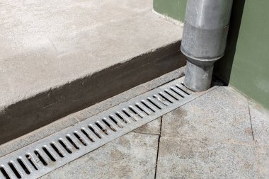 Mühendislik yapısı ön cephedeki borudan aşağı gri taş zeminin drenaj ızgarası ve çimento eşiği ile şehir altyapısının detaylarını kapatıyor..