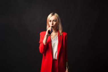 Genç beyaz sarışın kız güzel bir ceket ve beyaz bluz ile sıkı takım elbise kamera ve duman vape bir siyah görünüyor parlak kırmızı arka plan izole