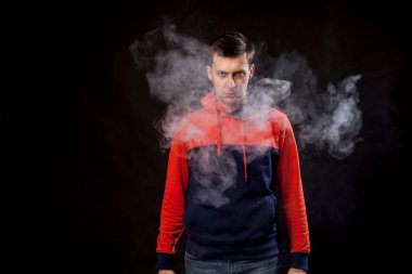 Genç bir adam bir portakal hoody ve onun fitness bilezik ile ayakta kot kol ve bir vape içiyor ve bir beyaz siyah bir izole arka plan üzerinde duman exhales