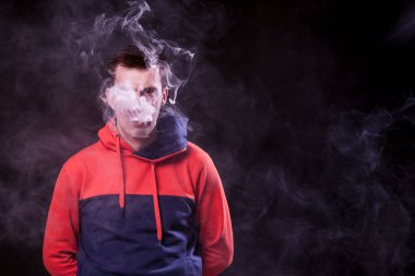 Hüzünlü gözleri renkli turuncu ve mavi kapüşonlu genç bir siyah saçlı adamla sigara arkasında silah ile bir vape duran ve bir beyaz siyah bir izole arka plan üzerinde duman exhales