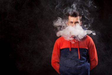 Hüzünlü gözleri renkli turuncu ve mavi kapüşonlu genç bir siyah saçlı adamla sigara arkasında silah ile bir vape duran ve bir beyaz siyah bir izole arka plan üzerinde duman exhales