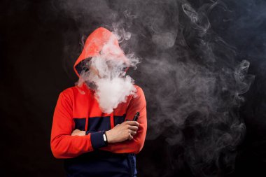 Genç esmer biri yakın duruş içinde renkli bir turuncu ve mavi kapüşonlu daimi bir vape içiyor ve bir beyaz siyah bir izole arka plan üzerinde duman exhales