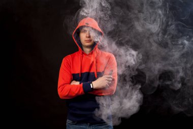 Genç esmer biri yakın duruş içinde renkli bir turuncu ve mavi kapüşonlu daimi bir vape içiyor ve bir beyaz siyah bir izole arka plan üzerinde duman exhales