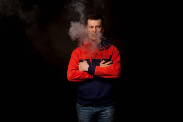 A young dark-haired man in a colored orange and blue hoody standing in close posture smokes a vape and exhales a white smoke on a black isolated background