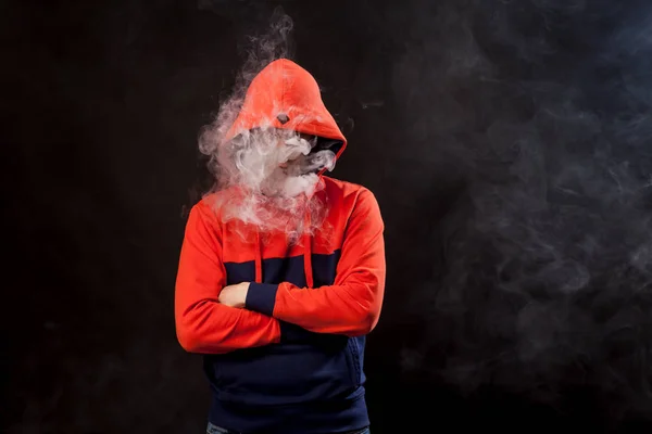 A young dark-haired man in a colored orange and blue hoody standing in close posture smokes a vape and exhales a white smoke on a black isolated background