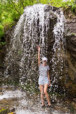 Genç sarışın bir kadın beyaz şapkalı mavi kot şort ve şelale altay dağlarında bir uçurumun üzerinde çizgili tişört duruyor