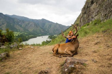 A young red-haired girl dressed in sports clothes stroking the horns and skin of a deer maral who lies on a mountain tied with a bridle in the mountains of the Altai clipart