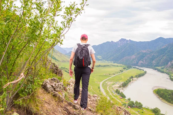 A young man in a cap with a backpack and black sports pants is going on the edge of a cliff in the mountains of the Altai, from where he opens a view of the river Katun and the island