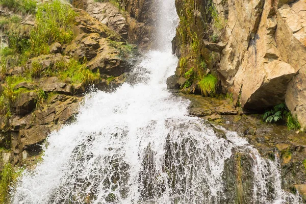 A high waterfall in the mountains of the Altai with sprinkled drops of water through rocks and rocks among green trees in the forest in sunny summer autumn weather
