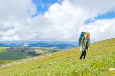Yeşil ceketli sarışın bir kız yalnız altay içinde dağın bir sırt çantası içinde belgili tanımlık geçmiş Vadisi, mavi gökyüzü ve yorgunluk ve bitkinlik hills doruklarına ile yüzünde yükselir.