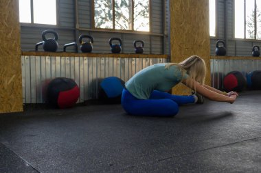 Sarışın kız uzun bir bacak için bükme yerde oturarak germe yapıyor, spor malzemeleri ile renkli ağırlık ve sağlık toplarıyla arka plan üzerinde spor salonunda İkinci bent