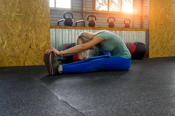 Sarışın kız üzerinde uzun bacaklarını bükme yerde oturarak germe onun ayak parmaklarını spor ekipmanları ağırlık ve sağlık toplarıyla arka plan üzerinde spor salonunda, tıraş yapıyor