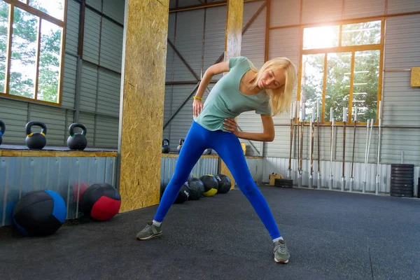 The blond girl in blue leggings in the gym is training, slanting slopes on the background of sports equipment: medical balls, dumbbells and bars, trees can be seen in the windows, the lights of a sun