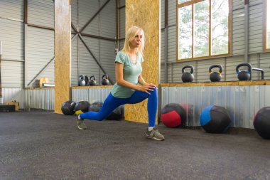 Genç göz kamaştırıcı olsun sarışın kız bacağını yükselterek kirpikleri tarafından germe yapıyor ve jimnastik salonu zemin karşı spor malzemeleri arka plan üzerinde yaslanmış, güneş pencereler arasında kırılıyor