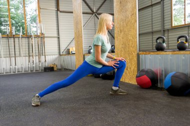 Sarışın güzel kadın bacağı yükselterek kirpikleri tarafından germe yapıyor ve jimnastik salonu zemin karşı spor malzemeleri arka plan üzerinde yaslanmış, güneş pencereler arasında kırılıyor