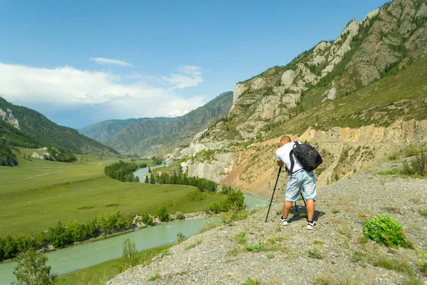Young man photographer travels through the Altai with the camera on the tripod takes a shot of the snow-covered mountains and the rocks with turquoise winding river Katun. Photo tour.