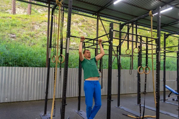 A young man in blue sports pants and a green T-shirt performs exercises pulling up on the horizontal bar in the gym outdoors with green grass on the background, rocky mountain, iron fenc