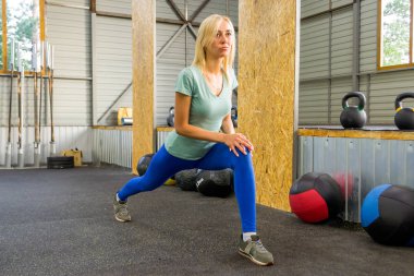 Sarışın güzel kız bacağını yükselterek kirpikleri tarafından germe yapıyor ve jimnastik salonu zemin karşı spor malzemeleri arka plan üzerinde yaslanmış, güneş pencereler arasında kırılıyor