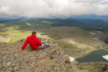 Genç bir adam çıplak ayakla ile bir dizüstü bilgisayar eller eserlerindeki serbest yazar olarak tehlikeli bir uçurumun dibinde olan yüksek bir dağda kenarına seyahat ederken oturur göl ve alan bitkiler ile