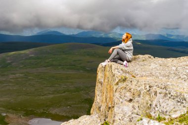 Güzel kızıl saçlı kadın oturur ve rahatlatıcı tutmak yalnız sakin bir rock Altay Dağları ile büyük gri bulutlar ve manzara mavi gökyüzü arka plan üzerinde ışık ışınlarının bir uçurumun kenarında