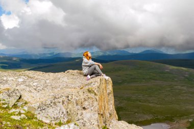 Güzel kızıl saçlı kız oturuyor ve rahatlatıcı sakin olsun yalnız bir rock Altay Dağları ile büyük gri bulutlar ve manzara mavi gökyüzü arka plan üzerinde ışık ışınlarının bir uçurumun kenarında