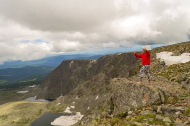 Kırmızı kapüşonlu genç bir adam bir dizüstü selfie sırasında tehlikeli bir uçurumun dibinde olan yüksek bir dağda kenarına seyahat ederken görüntülü sohbet yapmak onun elinde tutan göl ve alan