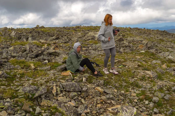 Two girls on the top of the mountain rest during the ascent of the mountain of the Altai in sport clothes with laptop in the arm on the background with a lot of stones and big clouds in the sky