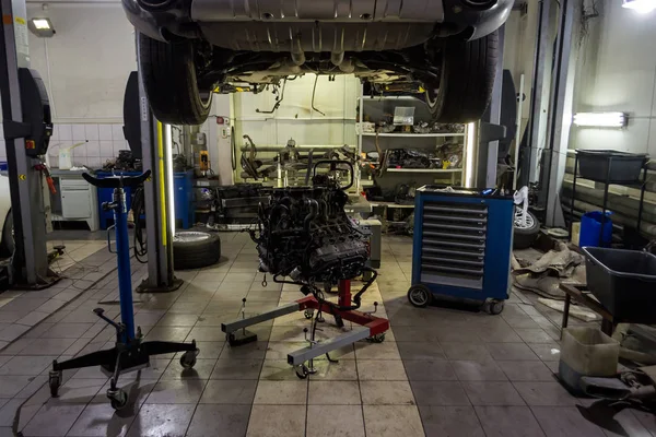 A sports car raised on a lift for repair and under it a detached engine suspended on a blue crane and a gear box on a lifting table in a vehicle repair shop with wheels lying on the sides