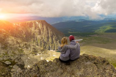 sevgi dolu bir çift bir kaya kenarında oturan onları önünde günbatımında bir ısmarlayarak dağlar, kar ile turkuaz göller Vadisi ve beyaz bulut ve güneş mavi bir gökyüzü kucaklayan