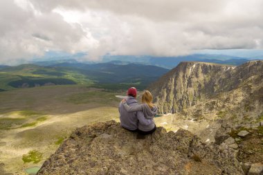sevgi dolu bir çift bir kaya kenarında oturan onları önünde bir ısmarlayarak dağlar, kar ile turkuaz göller Vadisi ve mavi gökyüzü beyaz bulutlar ile kucaklayan. Arkadan görünüş beni izleyin