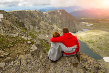 sevgi dolu bir çift bir kaya kenarında oturan onları önünde günbatımında bir ısmarlayarak dağlar, kar ile turkuaz göller Vadisi ve beyaz bulut ve güneş mavi bir gökyüzü kucaklayan
